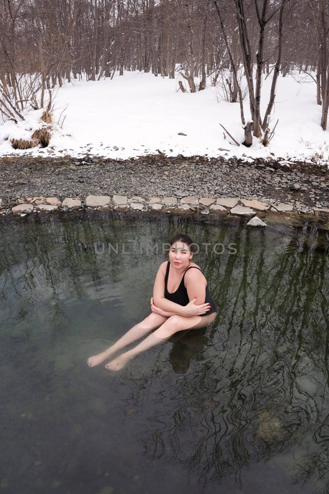 Large size young adult model in black one-piece bathing swimsuit sitting in geothermal mineral water in outdoors pool at balneotherapy spa, hot springs resort. Woman takes winter bathing and hardening