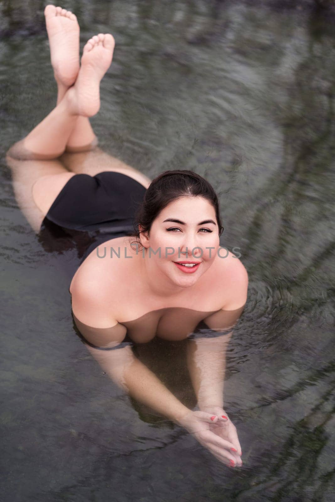 Curvy full figured model in black swimming costume lying in geothermal mineral water in outdoor pool at health spa, hot springs resort. Woman looking at camera, smiling. Body positive. High angle shot