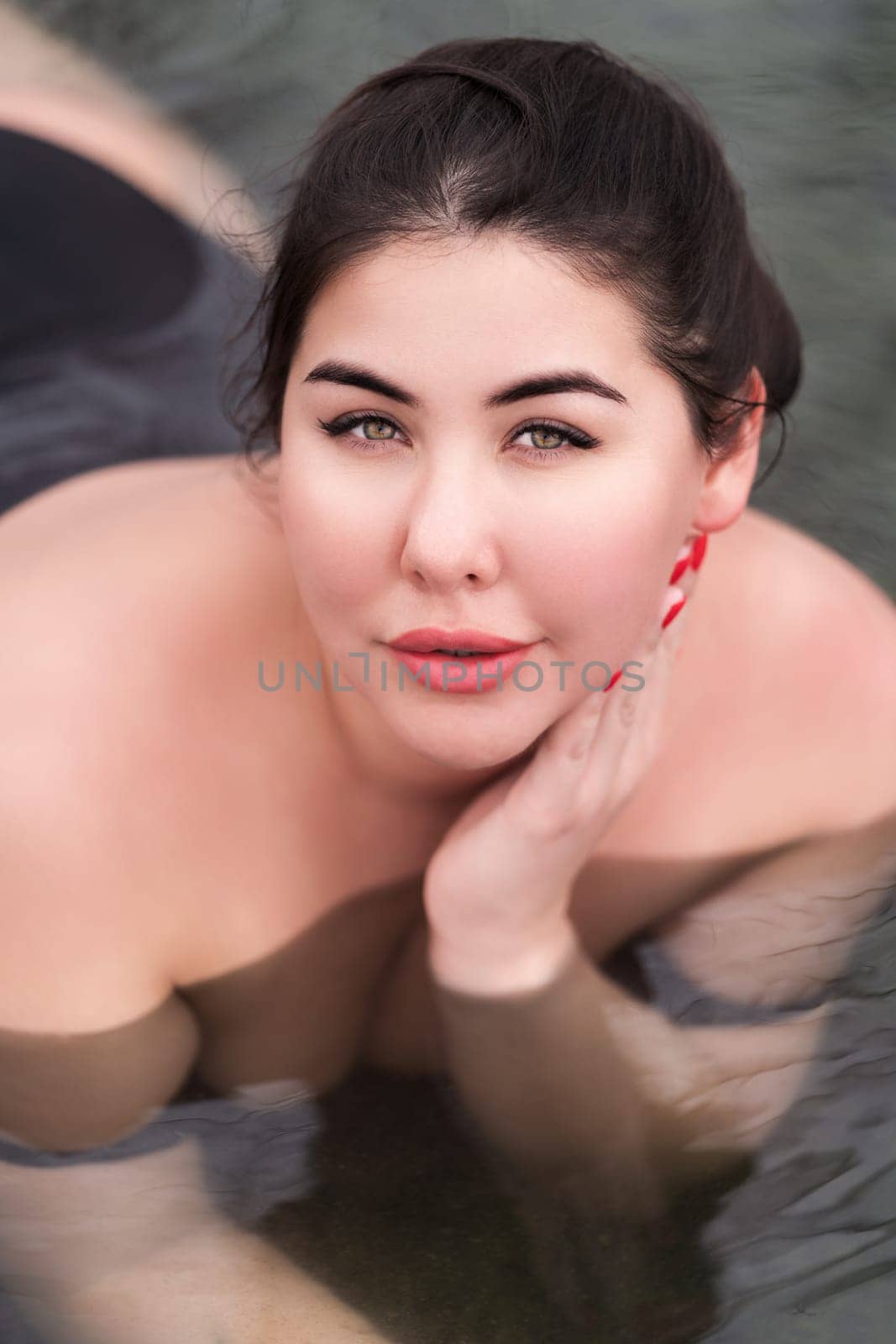 Portrait of brunette xxl young woman in black swimsuit relaxation lying down in outdoor pool at balneotherapy spa, hot springs resort. Authentic outsize young woman looking at camera. High angle shot