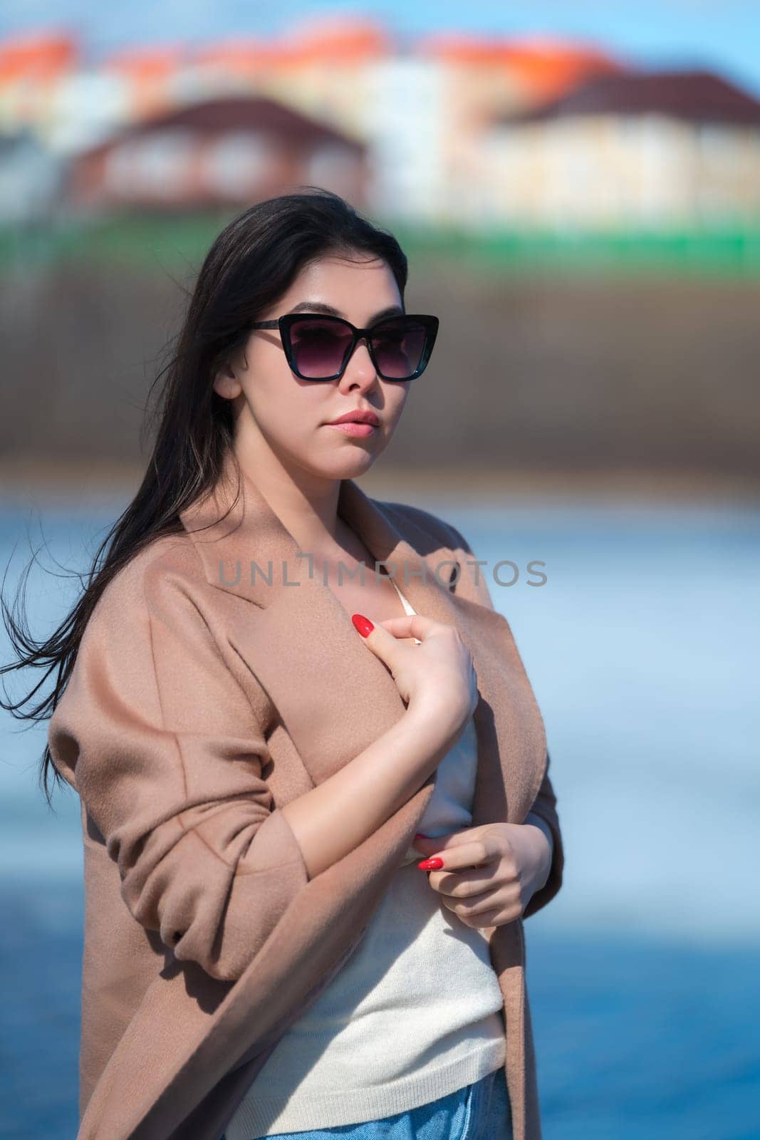 Portrait stylish young woman with long hair, plump lips and sunglasses, dressed in beige coat and white sweater posing outdoors against backdrop of blue lake and real estate standing in sunny weather.