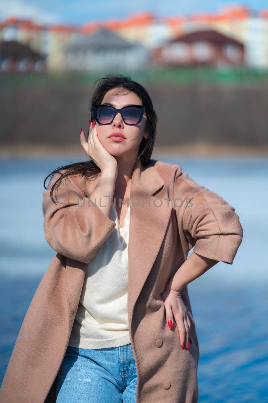 Sexy plus size young female with long hair, plump lips and sunglasses, dressed in beige coat, white sweater and blue jeans posing outdoors against background of blue lake and real estate in sunny day