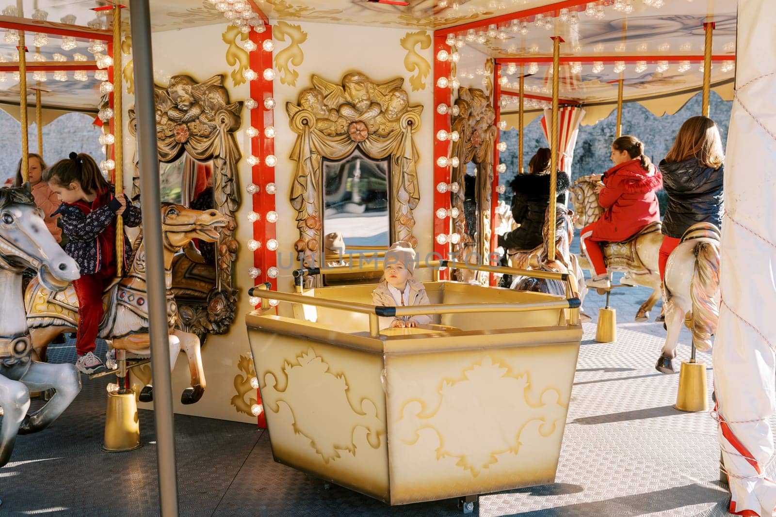 Little girl sits on a carousel seat next to children riding toy horses. High quality photo
