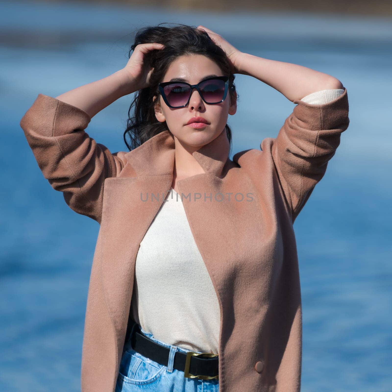 Fashionable young female with big breasts, long hair, plump lips and sunglasses, dressed in beige coat, white sweater and blue jeans posing outdoors against background of blue lake in sunny weather