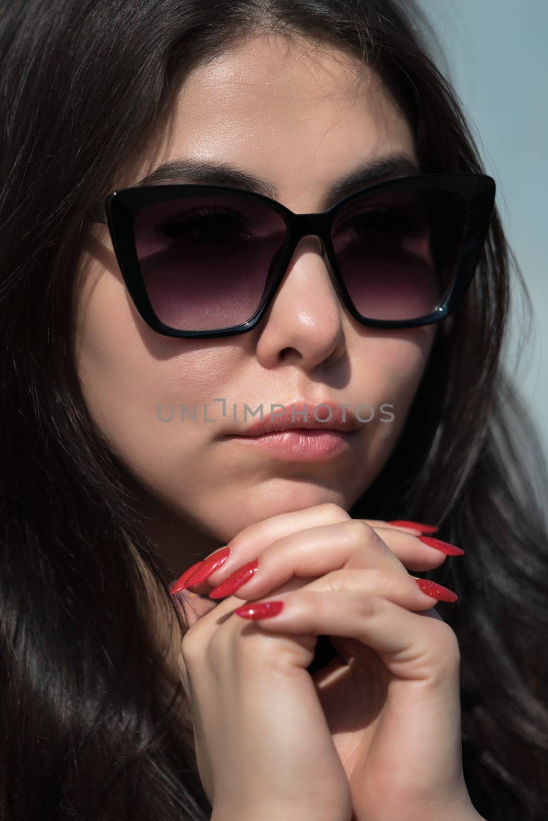 Close-up portrait of beauty young female model face with plump lips and glasses folded hands under her chin. Charming hipster young woman with black long hair looking at camera through dark sunglasses