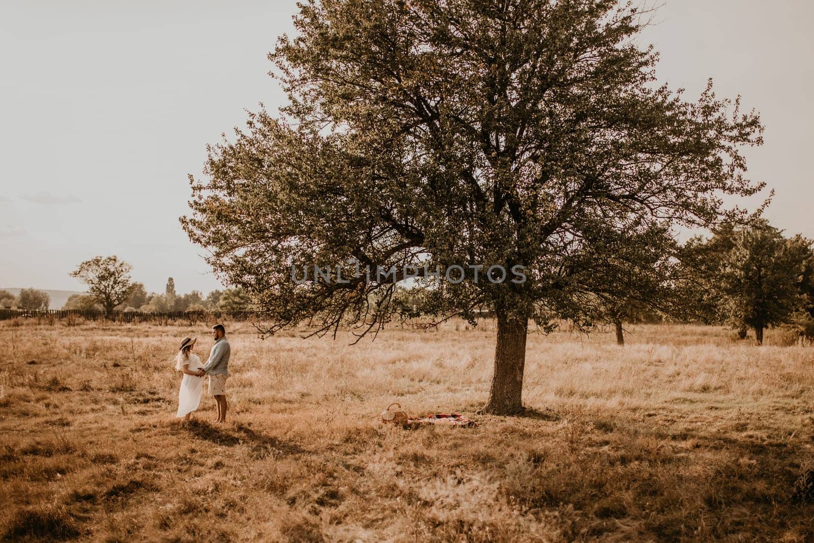 Happy family pregnant caucasian blonde woman in hat in white cotton dress walks with husband in meadow in summer. man in light natural clothes and shorts holds hand wife. trees in sunbeams
