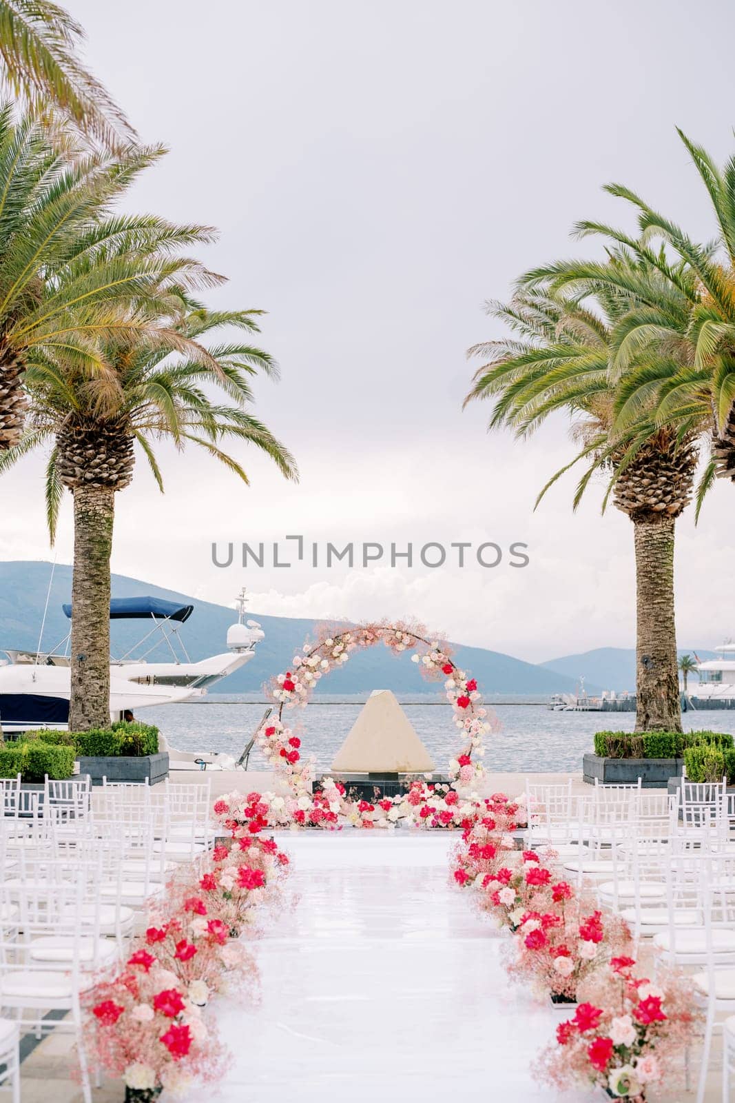Rows of chairs line a white path decorated with red flowers in front of a round wedding arch on the pier by Nadtochiy