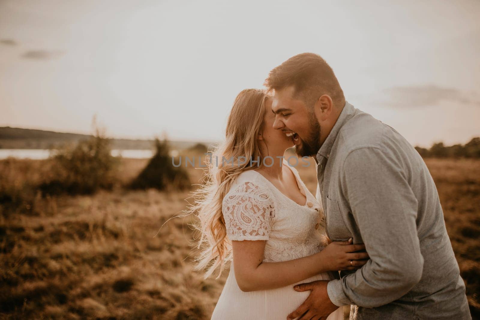 husband laughs hugs pregnant wife keep his hands on big round tummy. Happy family resting in nature hugs kisses in summer at sunset. future mother Caucasian woman in white cotton dress father