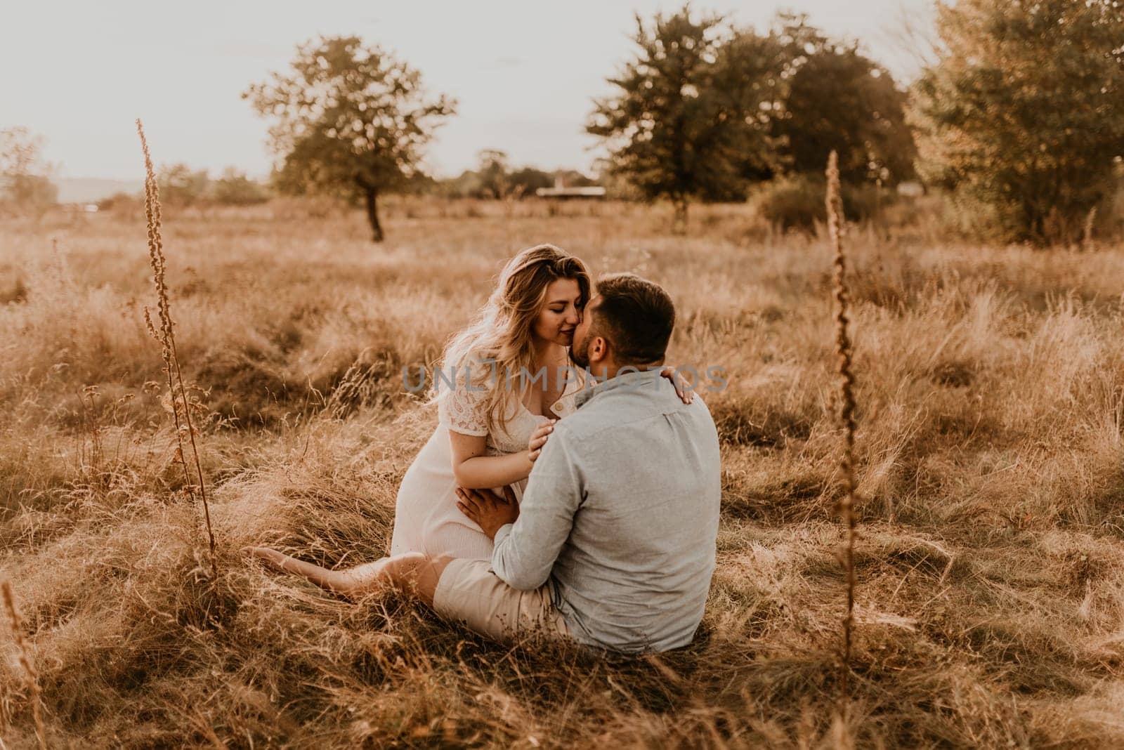 pregnant wife sits in her husband arms and kisses and hugs him. Happy family resting nature hugs kisses in summer at sunset. future mother Caucasian woman white cotton dress father in shorts