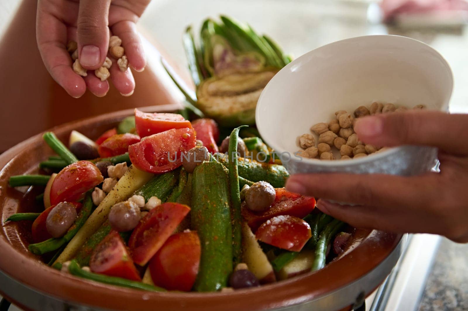 Closeup of the hands of a housewife adding chickpeas into a vegetarian meal while cooking healthy organic veggies in clay tagine pot. Traditional Moroccan food background