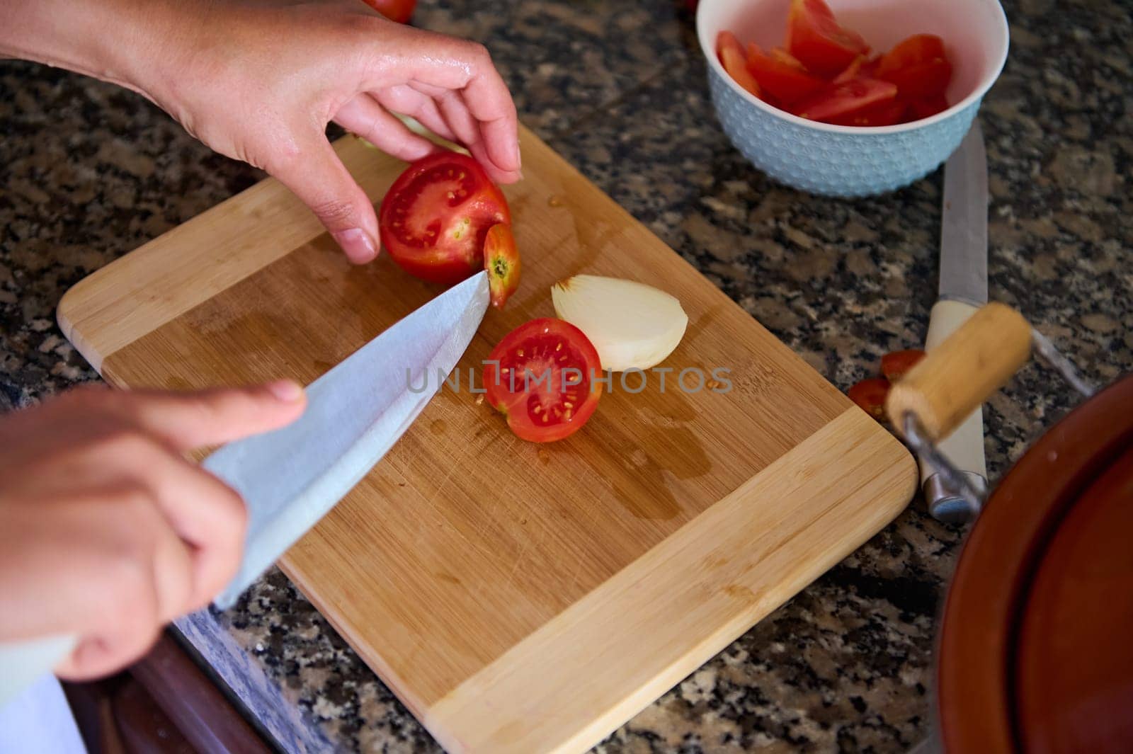 Top view of chef's hands using a kitchen knife, cutting fresh ripe organic tomatoes on a wooden board. Cook prepares a healthy salad