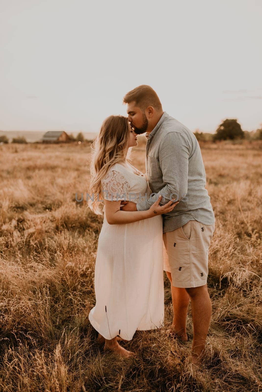 husband hugs pregnant wife keep his hands on big round tummy. Happy family resting in nature hugs kisses in summer at sunset. future mother Caucasian woman in white cotton dress father in shorts