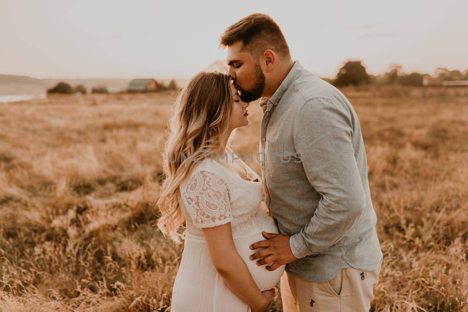 husband hugs pregnant wife keep his hands on big round tummy. Happy family resting in nature hugs kisses in summer at sunset. future mother Caucasian woman in white cotton dress father in shorts