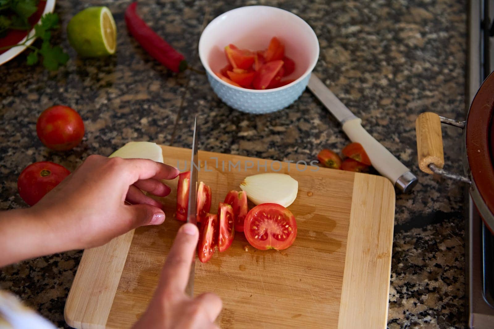 Close-up view from above of a housewife chopping tomatoes on a cutting board, cooking healthy raw vegan salad. Ingredients lying on a marble kitchen counter
