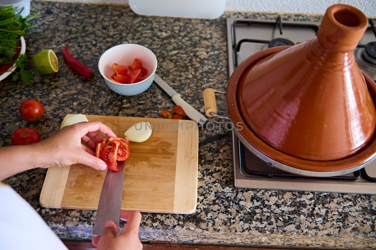 Close-up top view of a housewife slicing fresh ripe tomatoes on a cutting board, cooking healthy raw vegan salad. Ingredients lying on a marble kitchen counter and clay tajine pot on a steel stove