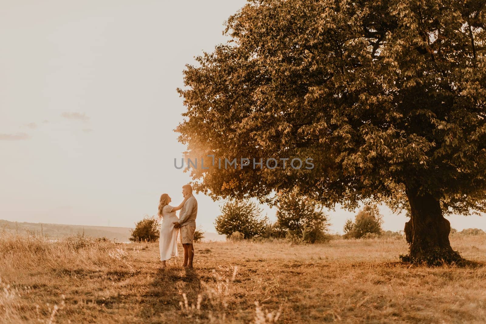 husband hugs pregnant wife with big tummy. Happy family resting in nature hugs kisses in summer at sunset. couple on background of large lonely tree. future mother Caucasian woman in cotton dress