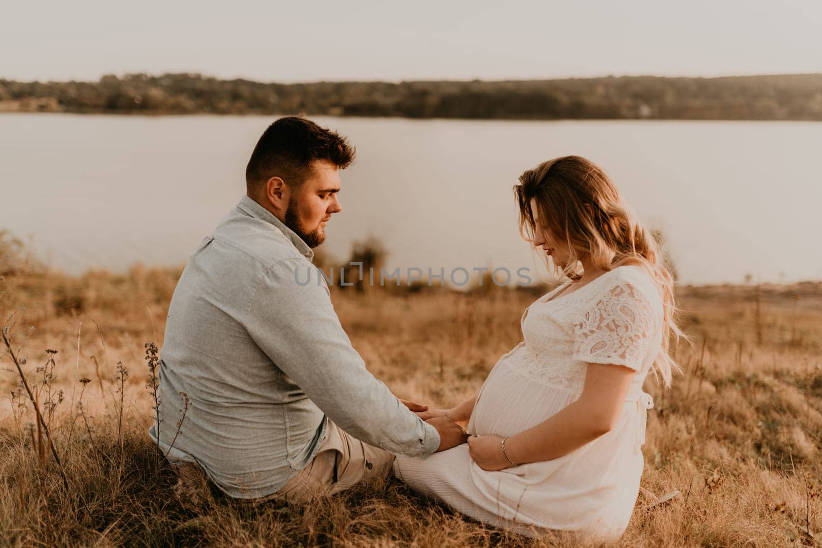 husband with pregnant wife with big belly tummy is resting on bank of river sitting on grass in summer sunny weather. couple in love caucasian man and woman hugging holding hands kissing outdoors