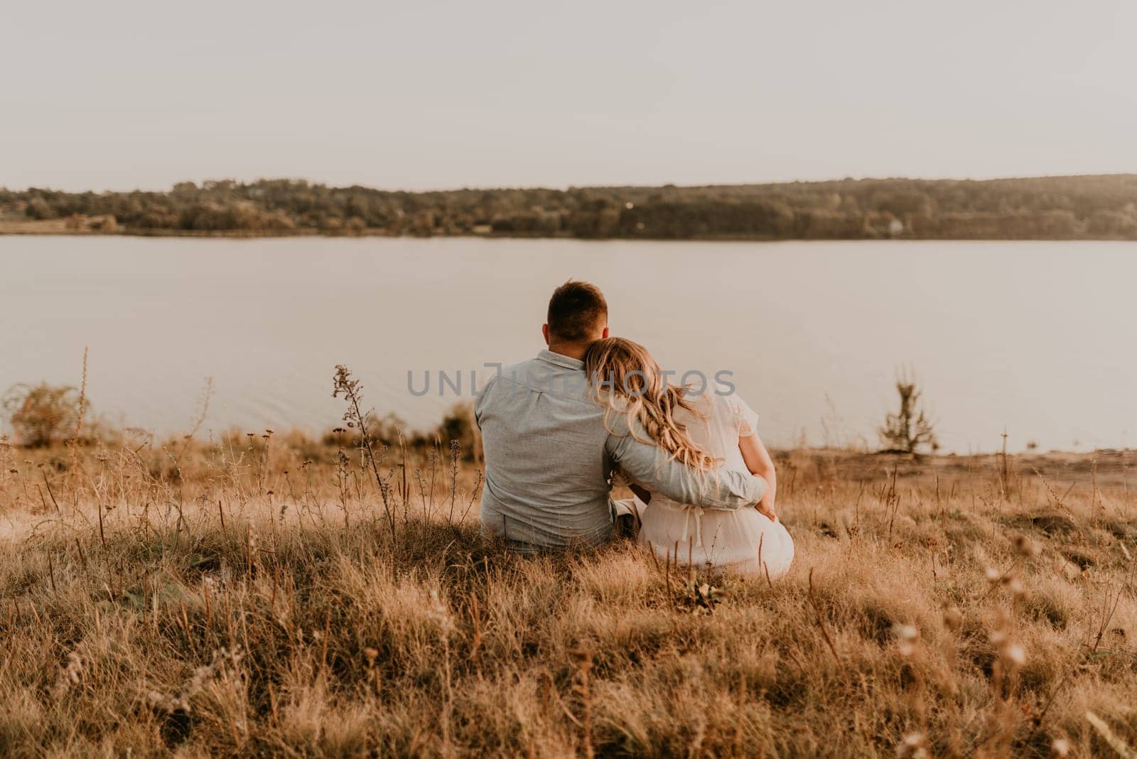 husband with pregnant wife with big belly tummy is resting on bank of river sitting on grass in summer sunny weather. couple in love caucasian man and woman hugging holding hands kissing outdoors