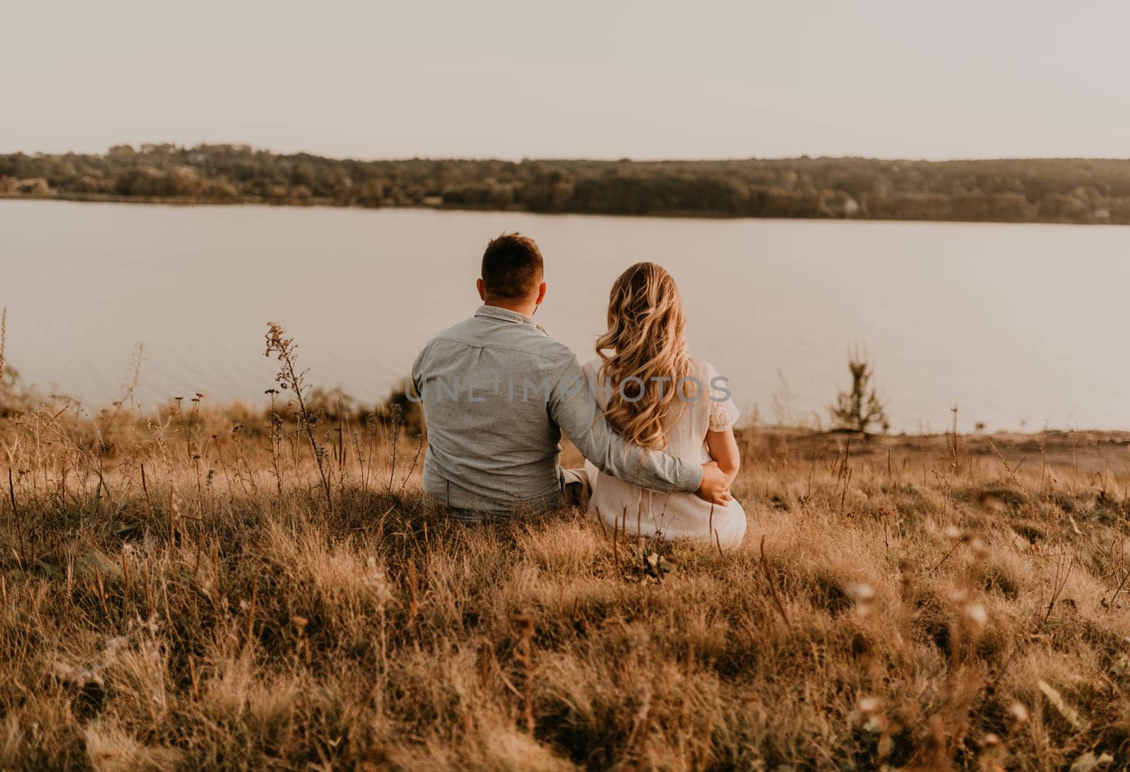 husband with pregnant wife with big belly tummy is resting on bank of river sitting on grass in summer sunny weather. couple in love caucasian man and woman hugging holding hands kissing outdoors