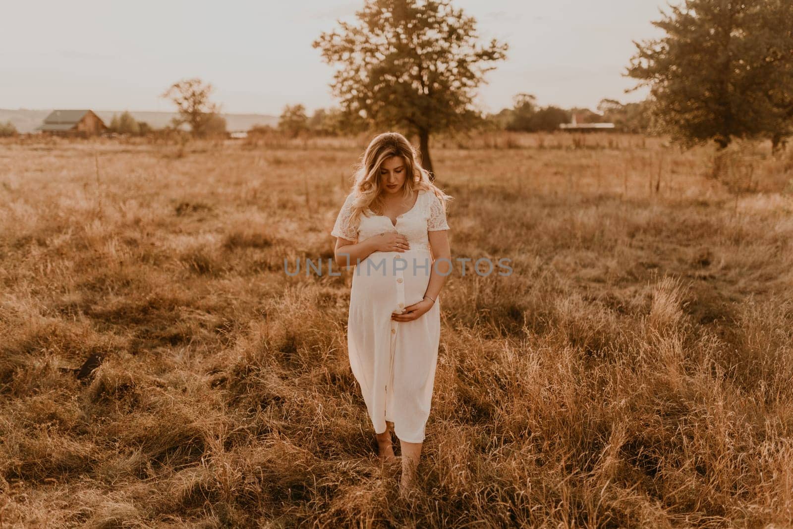 Caucasian pregnant young blonde woman in cotton white linen dress stand walking meadow on dry grass in summer at sunset nature. mother-to-be holds her hands on big round belly. tree in sunbeam