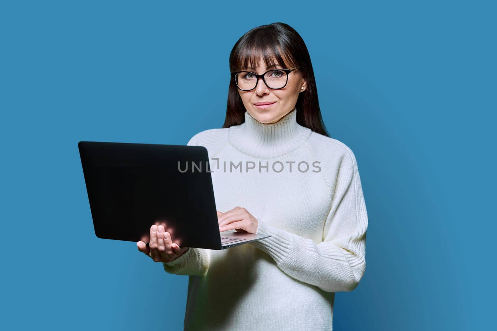 Middle-aged beautiful woman holding laptop looking at camera on blue studio background. Smiling happy female in white sweater using computer. Technologies for business work communications leisure