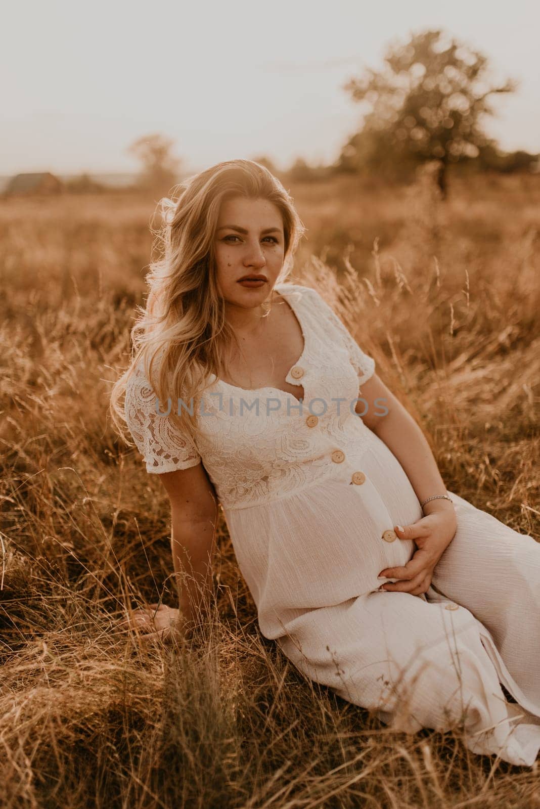 Caucasian pregnant young blonde woman in cotton white linen dress sits and lies in the middle of the meadow on dry grass in summer at sunset in nature. mother-to-be holds her hands on big round belly