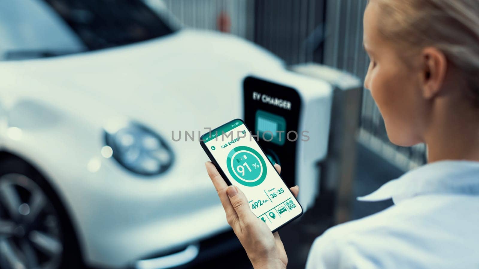 Businesswoman holding smartphone display battery status interface by smart EV mobile application while EV car recharging electricity from charging station in car park. Peruse