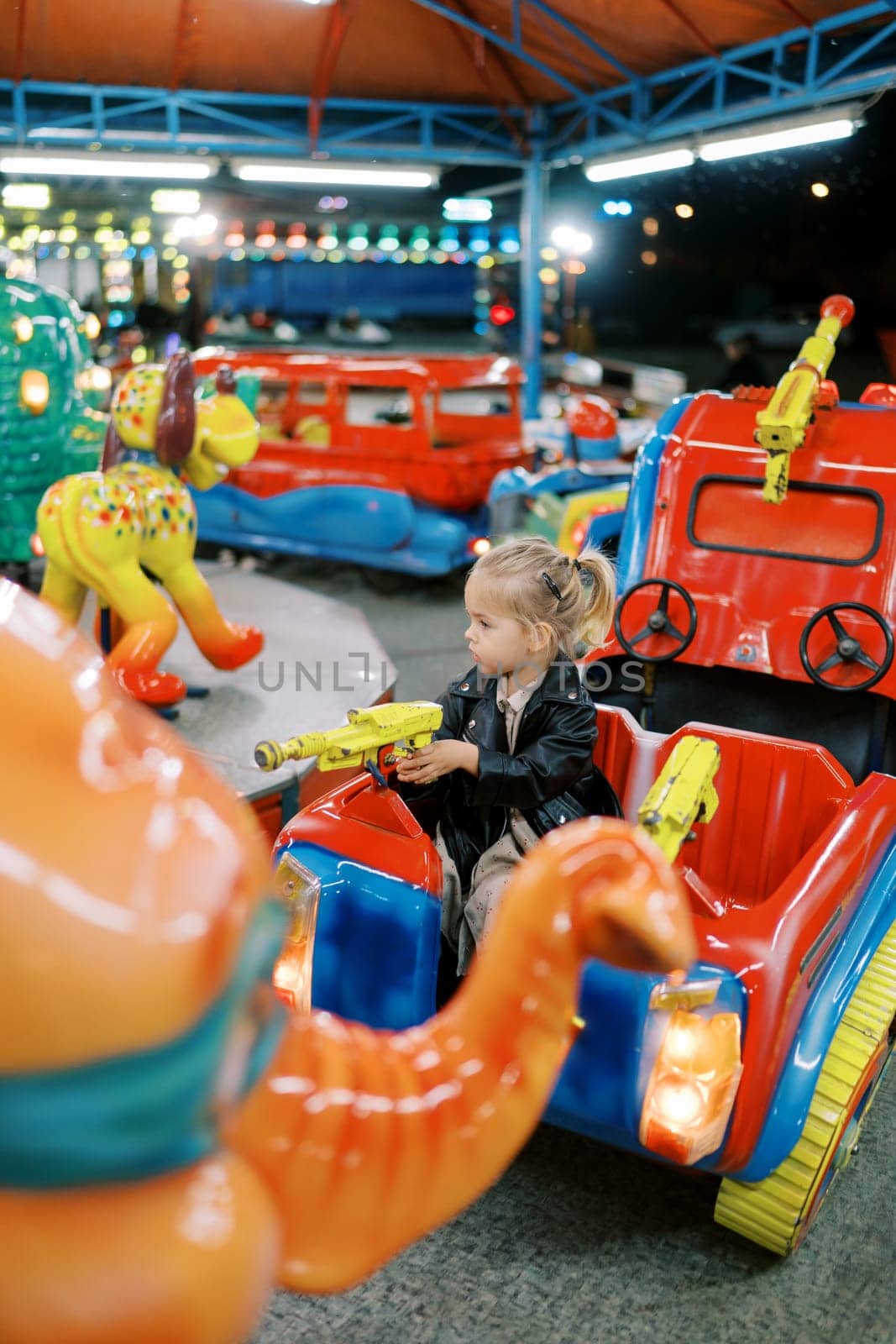 Little girl sits in a toy car on a carousel, holding the pistol steering wheel and looking to the side by Nadtochiy