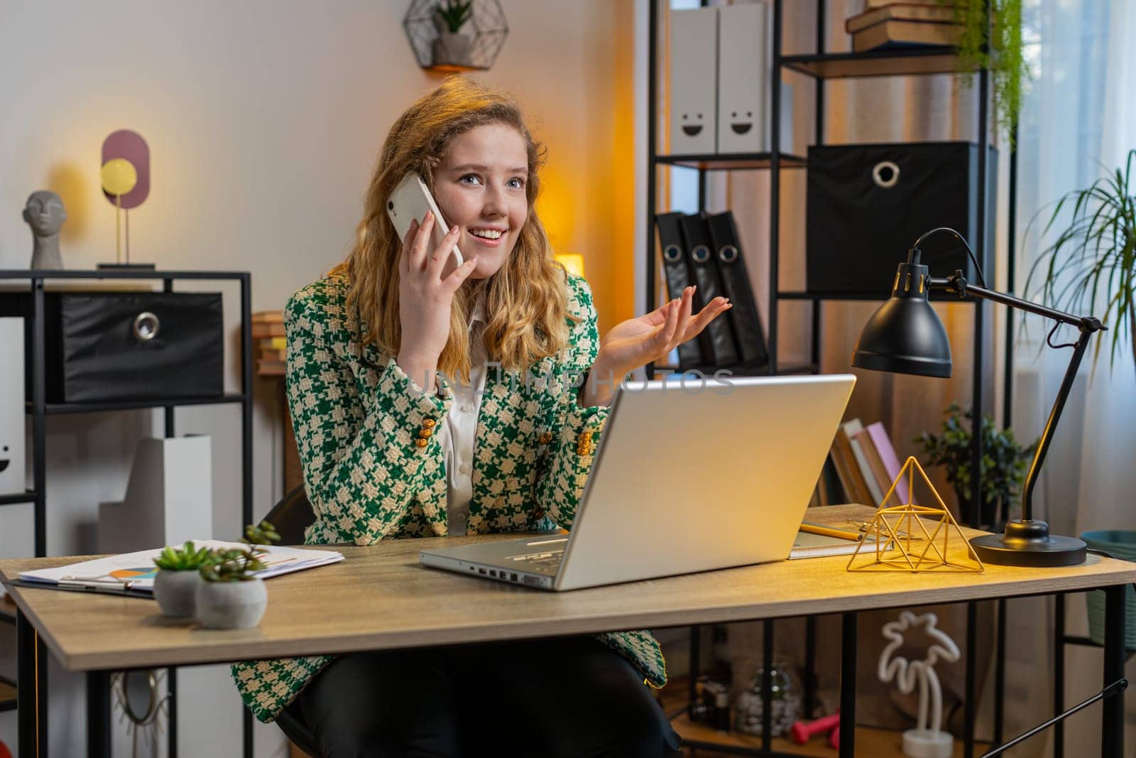 Young businesswoman freelancer answering to client on mobile phone call, using smartphone and laptop. Conversation. Caucasian woman girl working online remote distance job talking at home office table