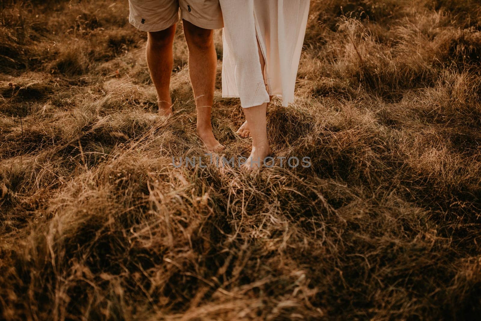 tanned man and light skin legs woman barefoot close-up walking on dry green thin grass. couple in love in summer in thin light cotton clothes walks in hot warm weather. women dress and men shorts