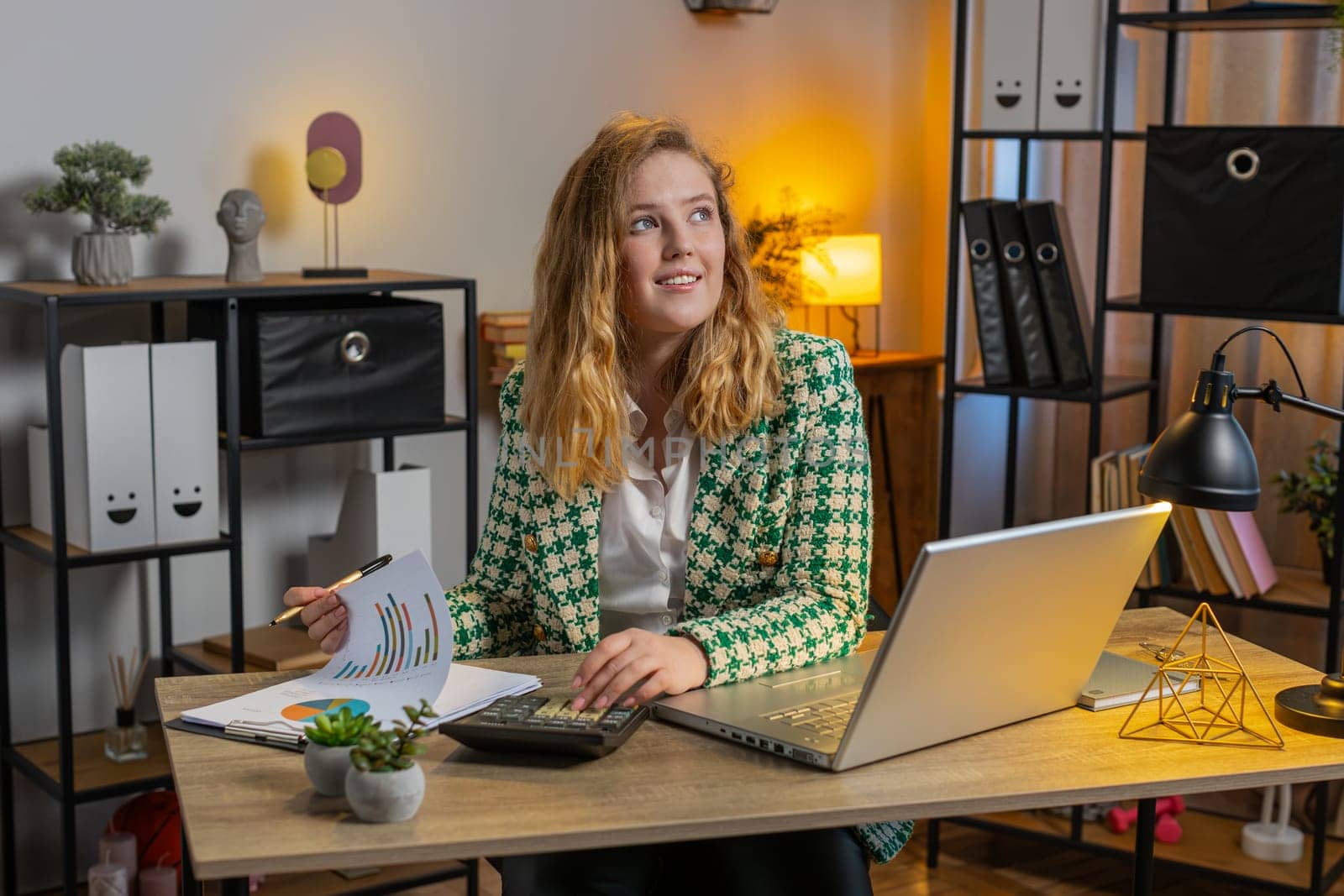 Happy smiling young freelancer analyzing paperwork graphs, accounting payments and calculating monthly expenses bank bills at home office desk. Blond businesswoman working online remote job on laptop