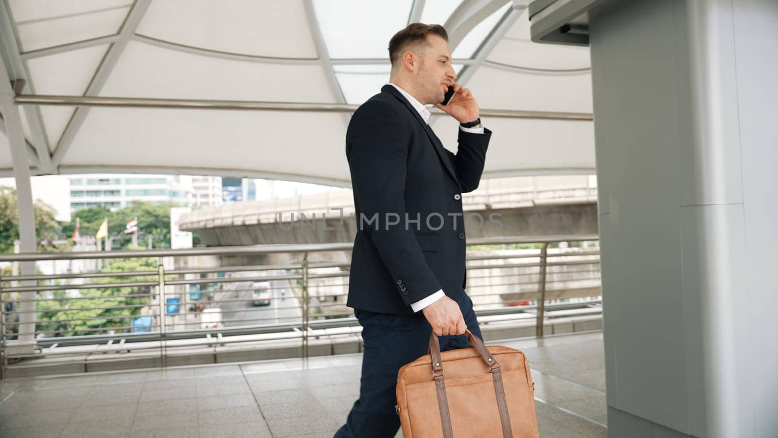 Smart caucasian businessman calling his colleague to plan financial strategy while walking to workplace. Side view of manager using mobile phone to communicate with marketing team. Lifestyle. Urbane.