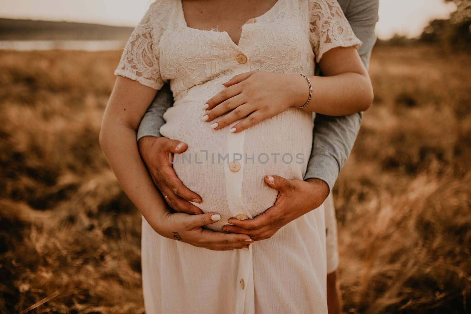 hands of future mother and father clasped pregnant big tummy. husband hugs pregnant wife. Happy family resting in nature hugs in summer at sunset. Caucasian woman in white cotton dress