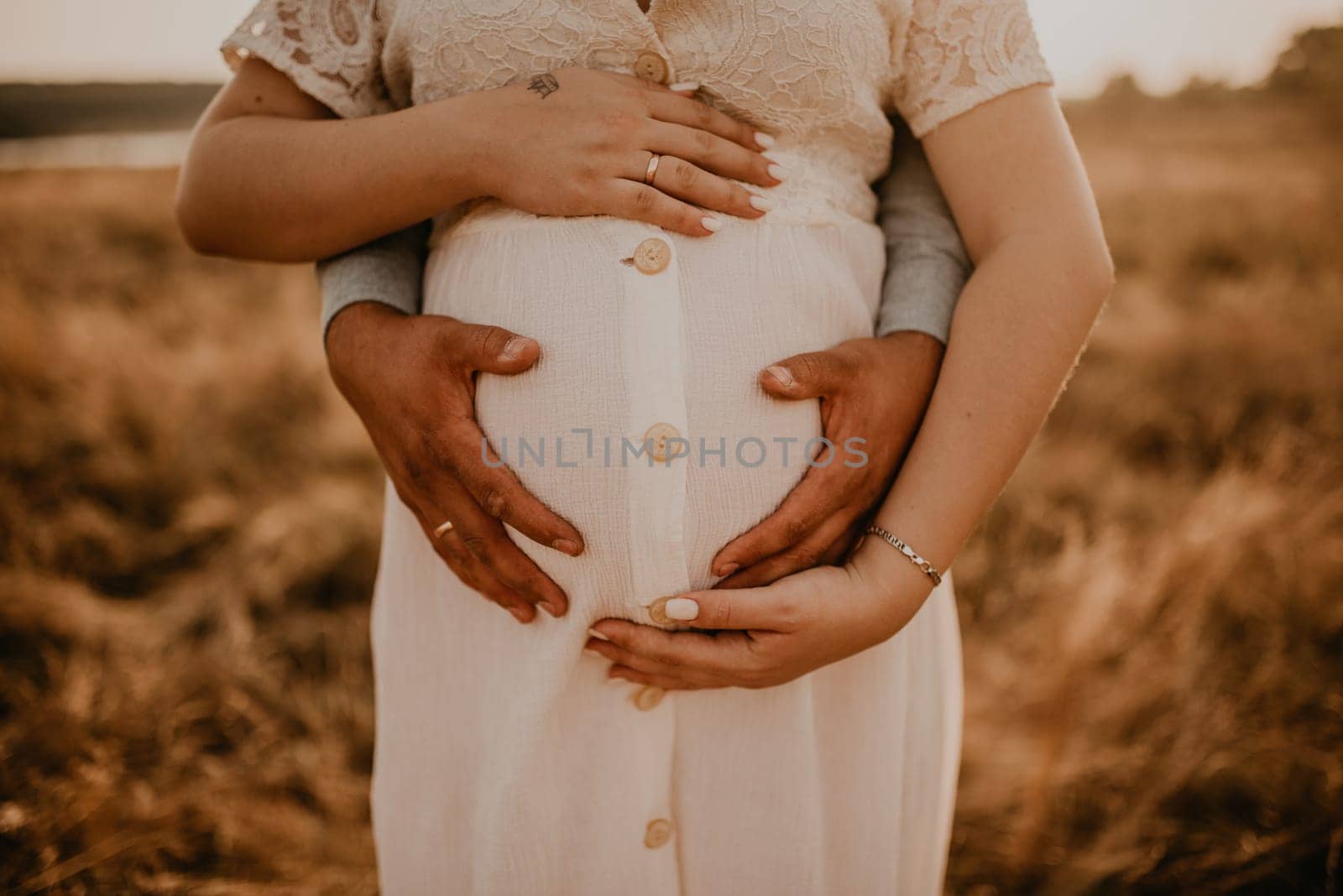 hands of future mother and father clasped pregnant big tummy. husband hugs pregnant wife. Happy family resting in nature hugs in summer at sunset. Caucasian woman in white cotton dress