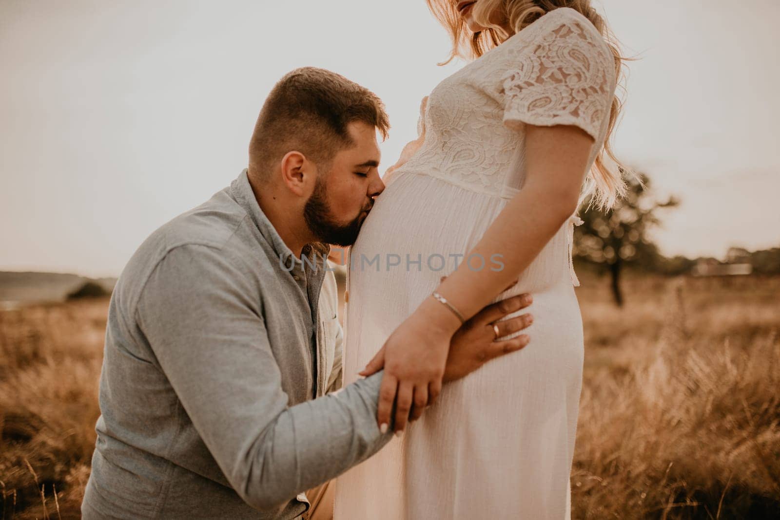 husband hugs pregnant wife hold hands on big round tummy kissing belly. Happy family resting in nature hugs kisses in summer at sunset. future mother Caucasian woman in white cotton dress. father