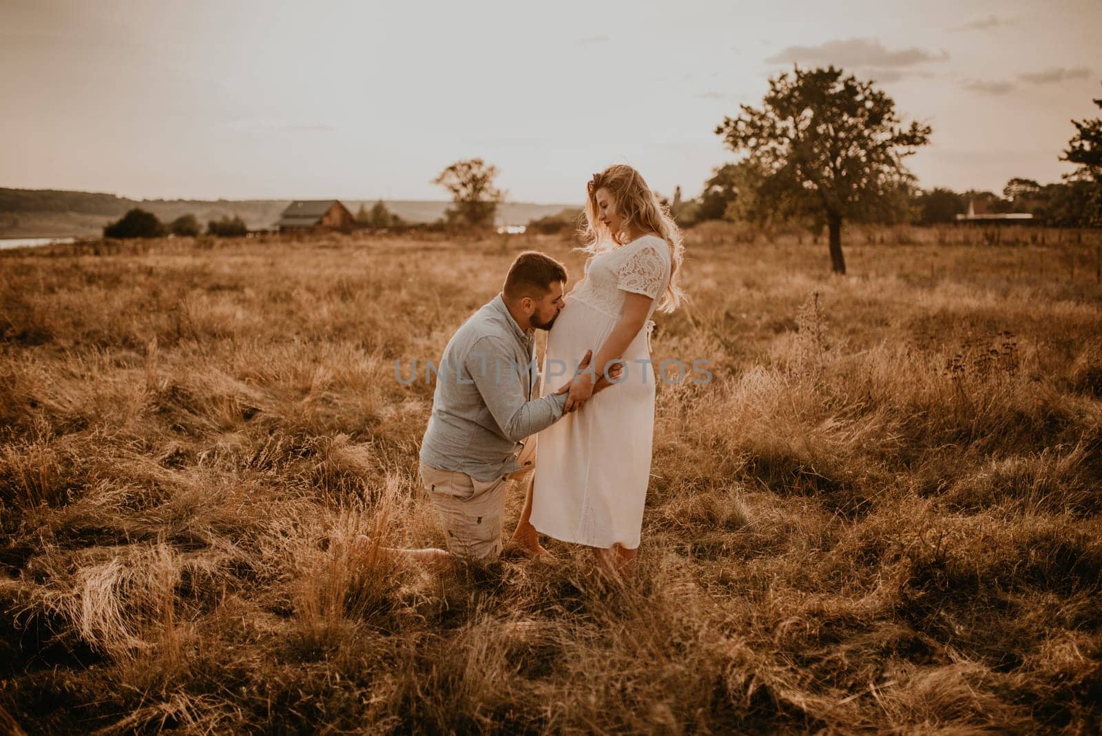husband hugs pregnant wife hold hands on big round tummy kissing belly. Happy family resting in nature hugs kisses in summer at sunset. future mother Caucasian woman in white cotton dress. father