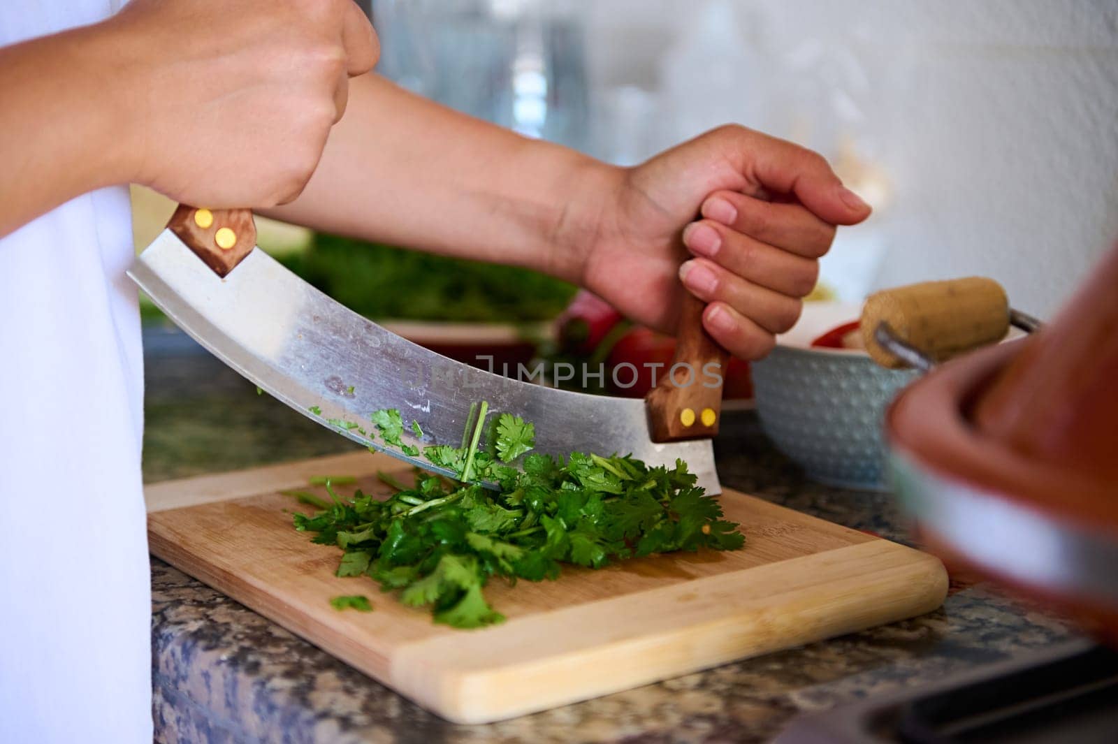 Close-up chef's hands using mezzaluna knife, chopping fresh green herb of parsley on a bamboo cutting board. Preparing healthy food.