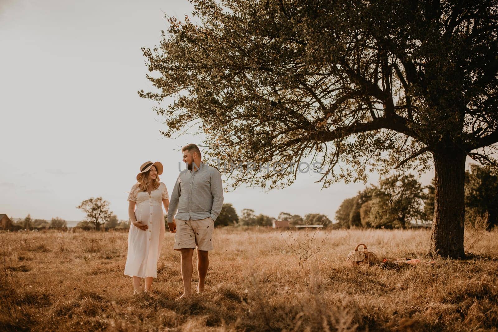 Happy family pregnant caucasian blonde woman in hat in white cotton dress walks with husband in meadow in summer. man in light natural clothes and shorts holds hand wife. trees in sunbeams