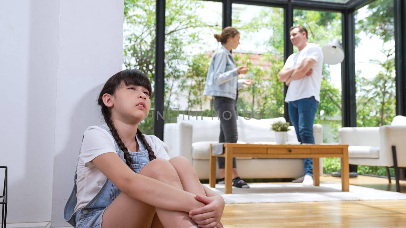 Stressed and unhappy young girl huddle in corner, cover her ears blocking sound of her parent arguing in background. Domestic violence at home and traumatic childhood develop to depression. Synchronos