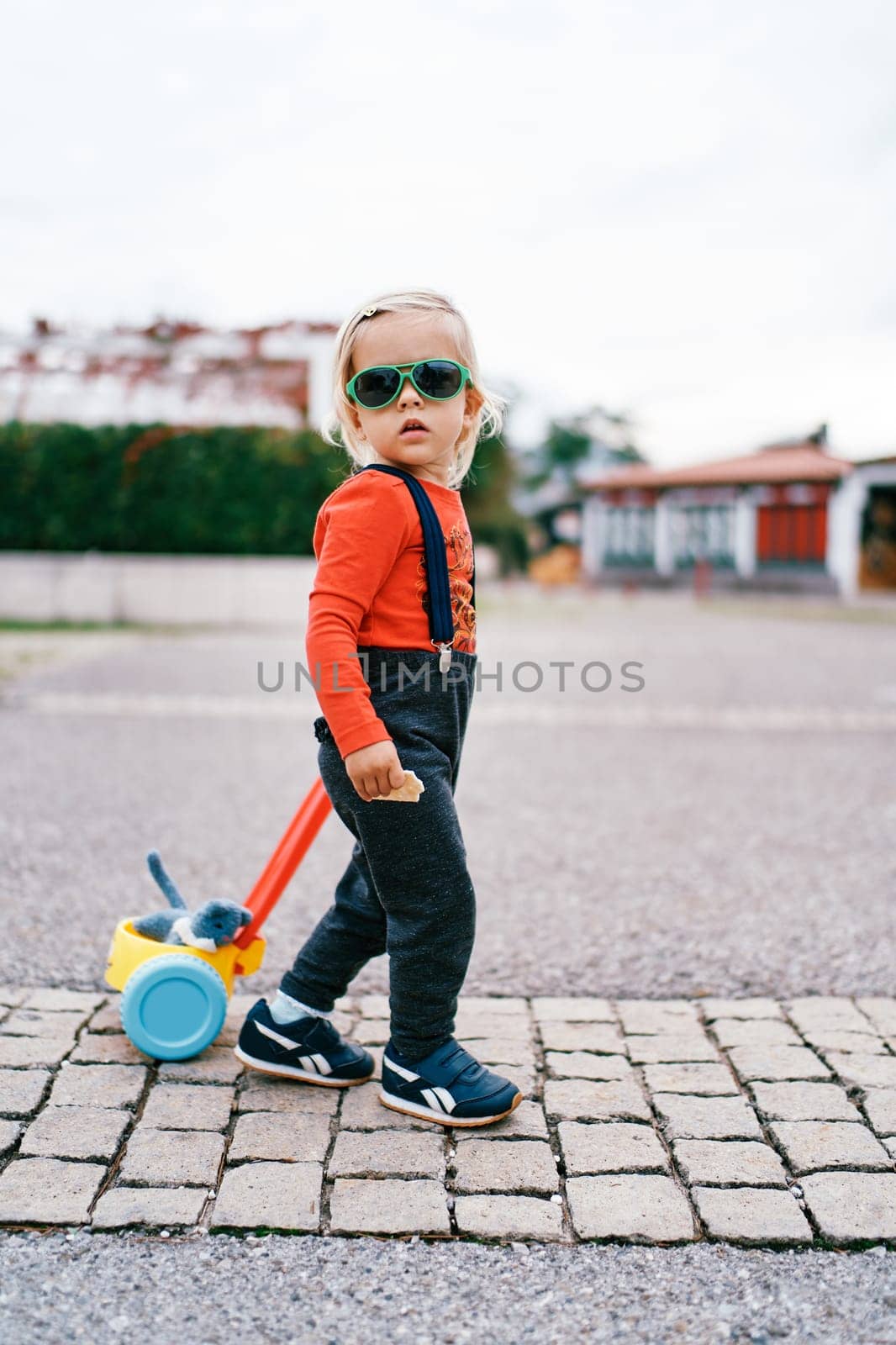 Little girl is pushing a toy cart along the paving stones, turning her head to the side by Nadtochiy