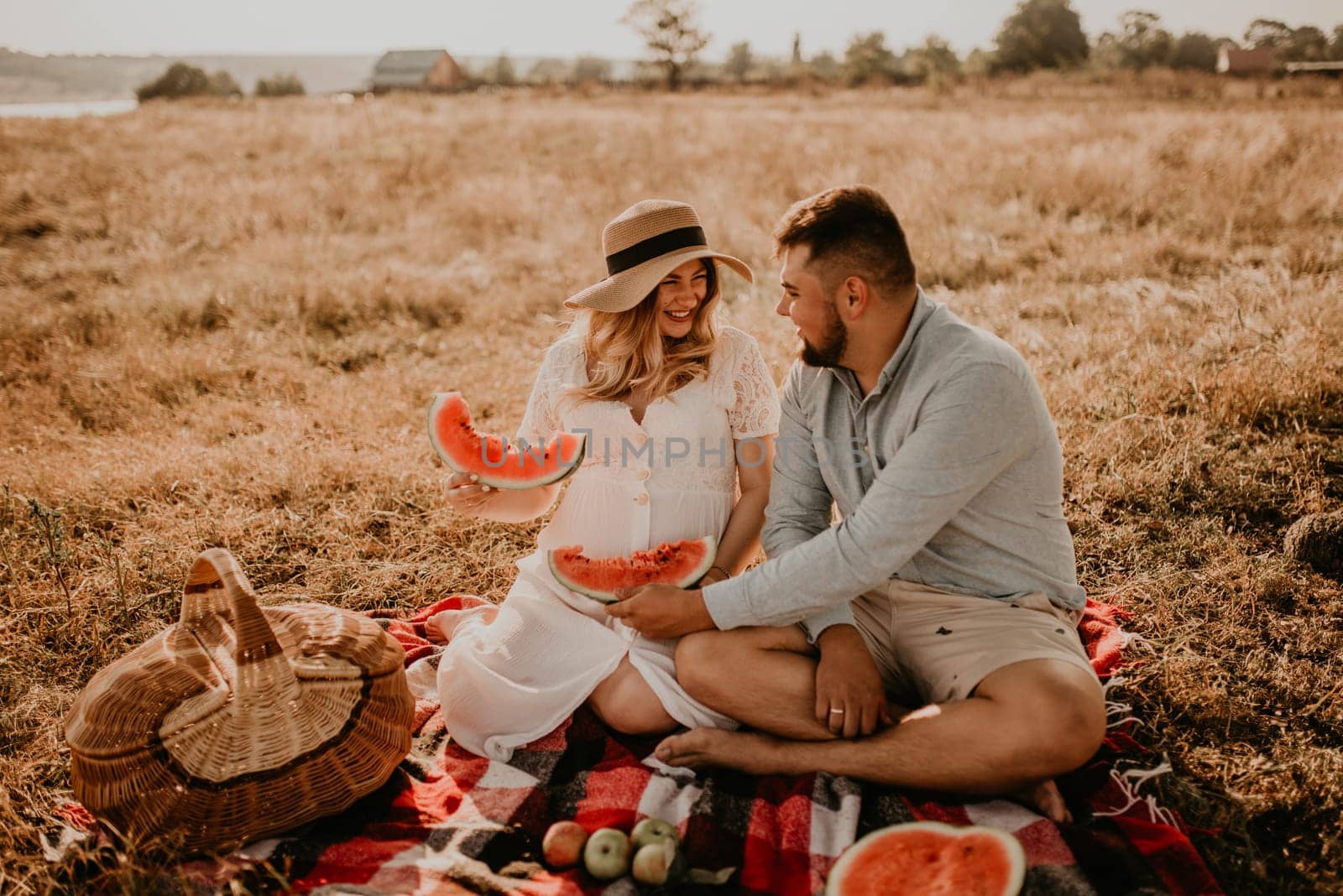 happy European caucasian family with a pregnant woman relaxing in nature picnic eating fruit red juicy watermelon laugh having fun. expectant mother in hat and dress eating watermelon in summer.