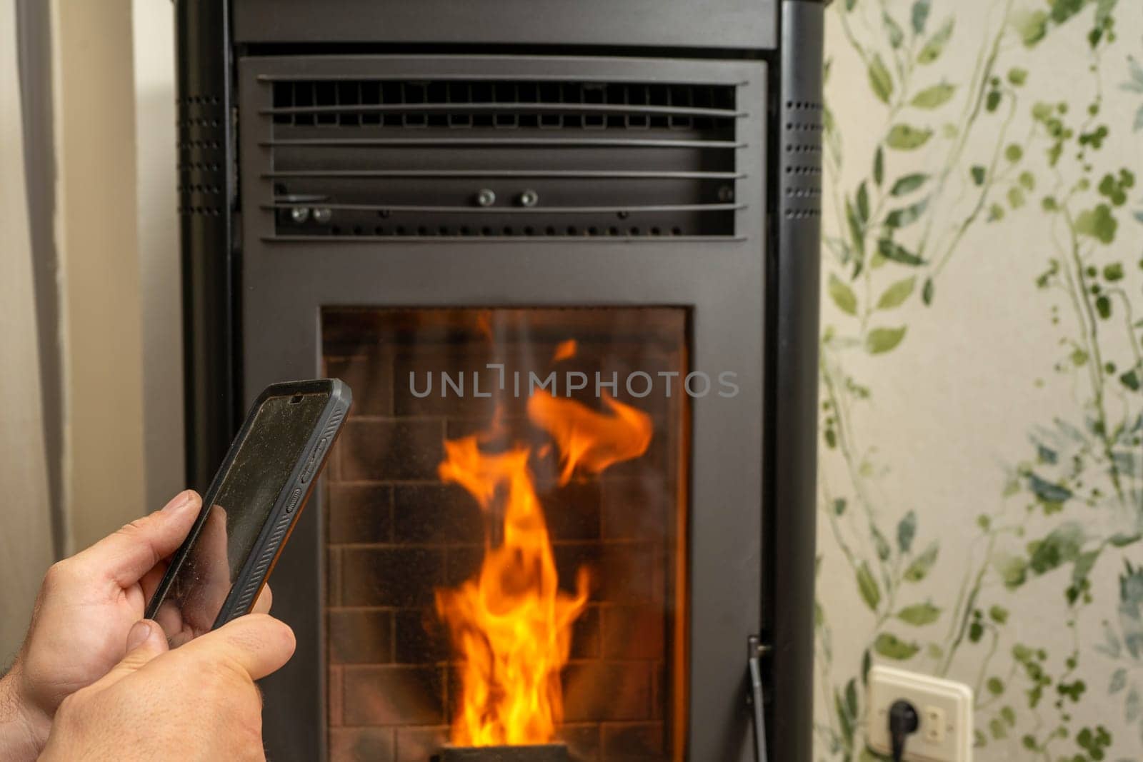 Man's hands with a cell phone controlling the modern pellet stove through its application, ecological heat. Sustainable energy