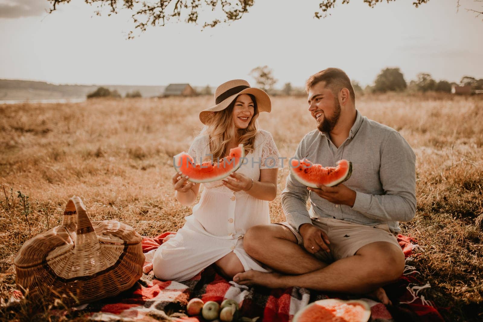 happy European caucasian family with a pregnant woman relaxing in nature picnic eating fruit red juicy watermelon laugh having fun. expectant mother in hat and dress eating watermelon in summer.