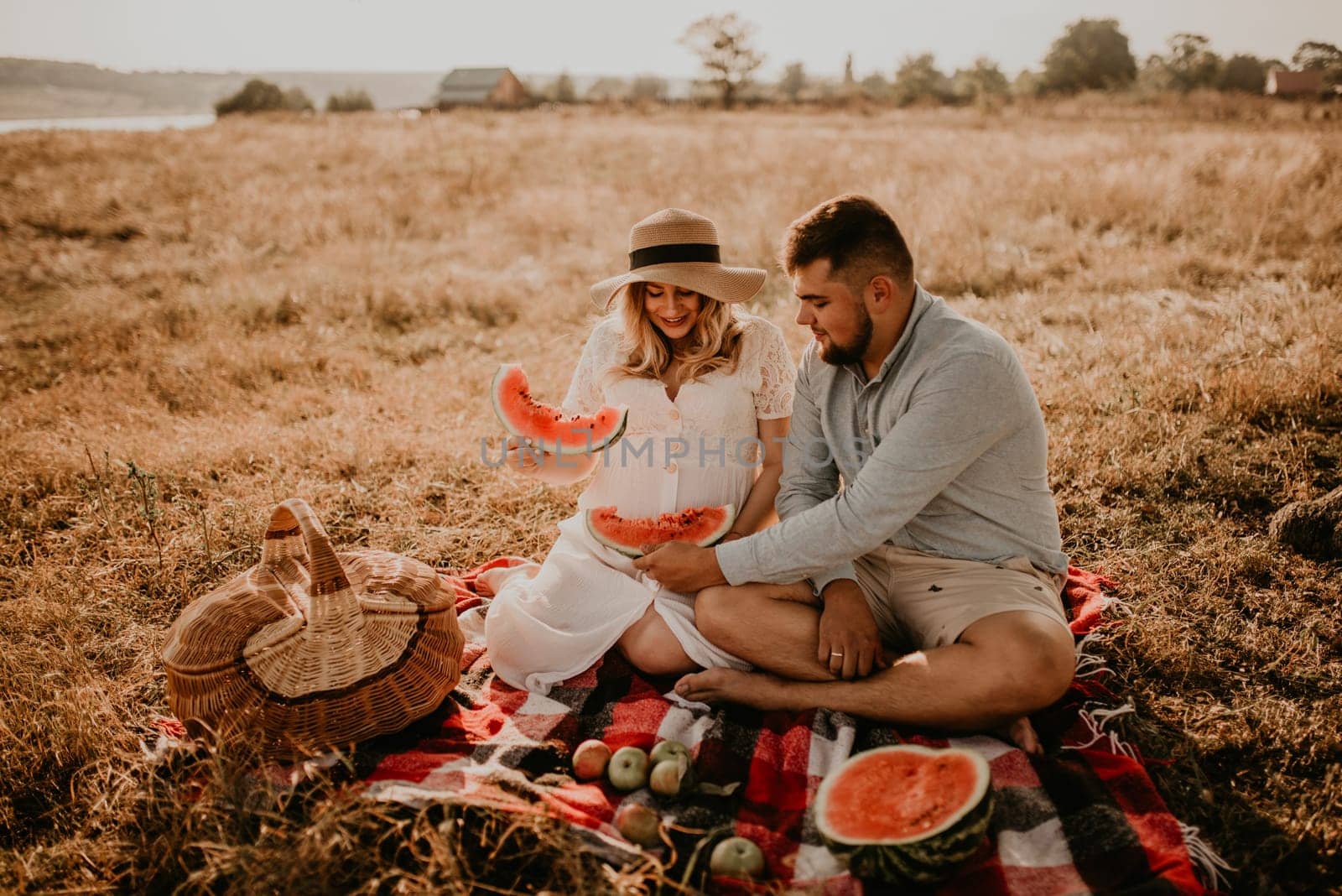 happy European caucasian family with a pregnant woman relaxing in nature picnic eating fruit red juicy watermelon laugh having fun. expectant mother in hat and dress eating watermelon in summer.