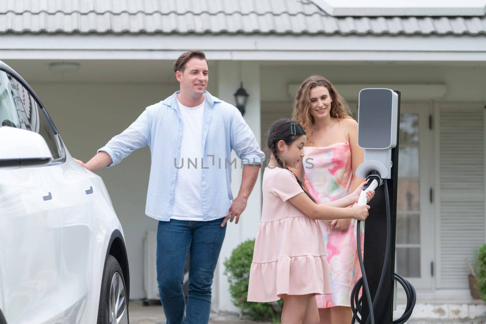 Happy little young girl learn about eco-friendly and energy sustainability as she help her family recharge electric vehicle from home EV charging station. EV car and modern family concept. Synchronos