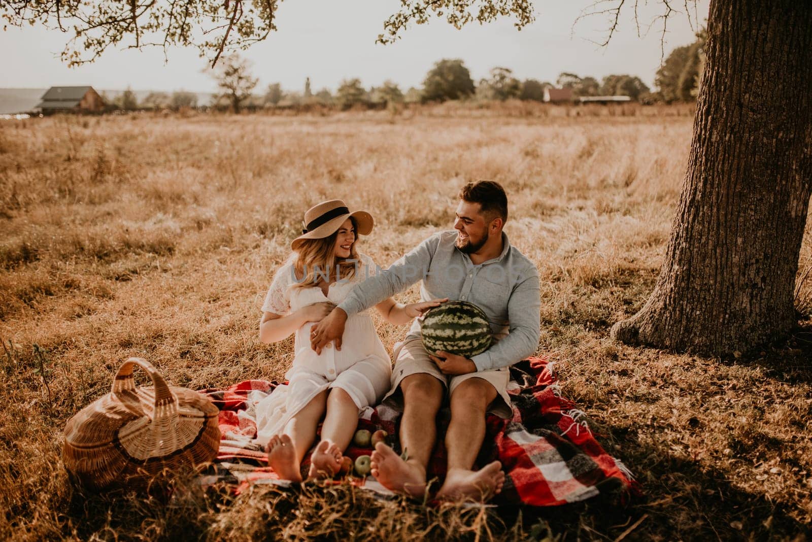 happy European caucasian family with a pregnant woman relaxing in nature picnic eating fruit red juicy watermelon laugh having fun. expectant mother in hat and dress eating watermelon in summer.