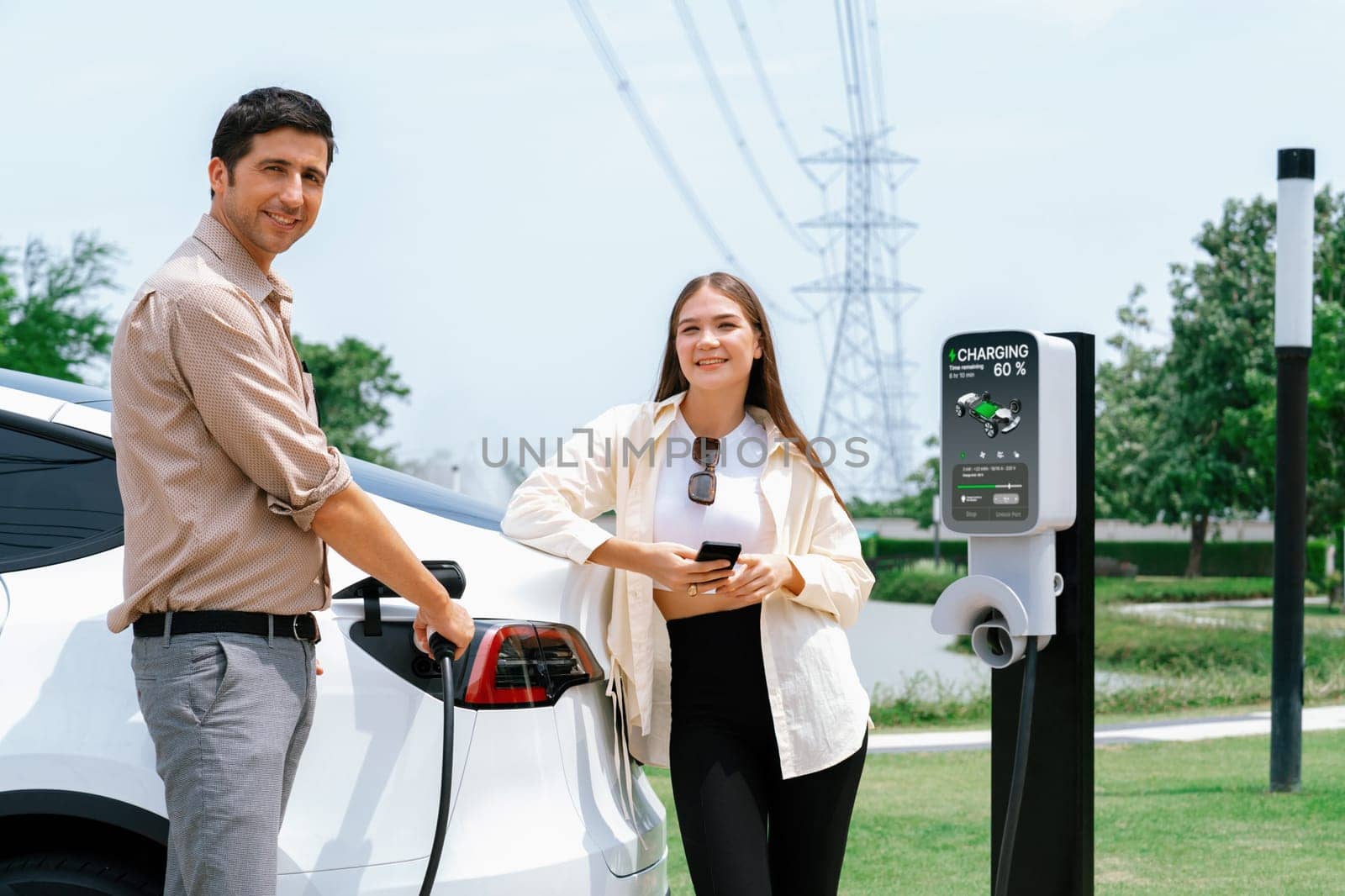 Young couple use smartphone to pay for electricity at public EV car charging station green city park. Modern environmental and sustainable urban lifestyle with EV vehicle. Expedient