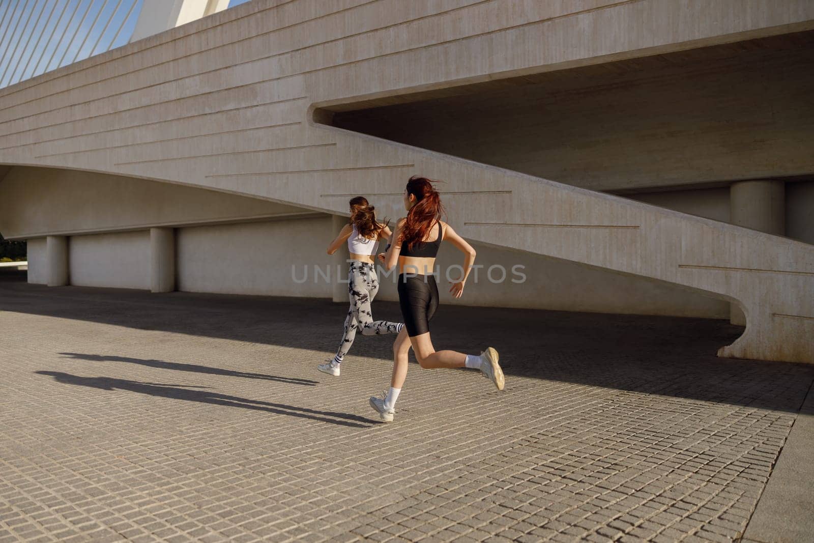Two young women in sportswear are running on modern buildings background. Active lifestyle concept by Yaroslav_astakhov