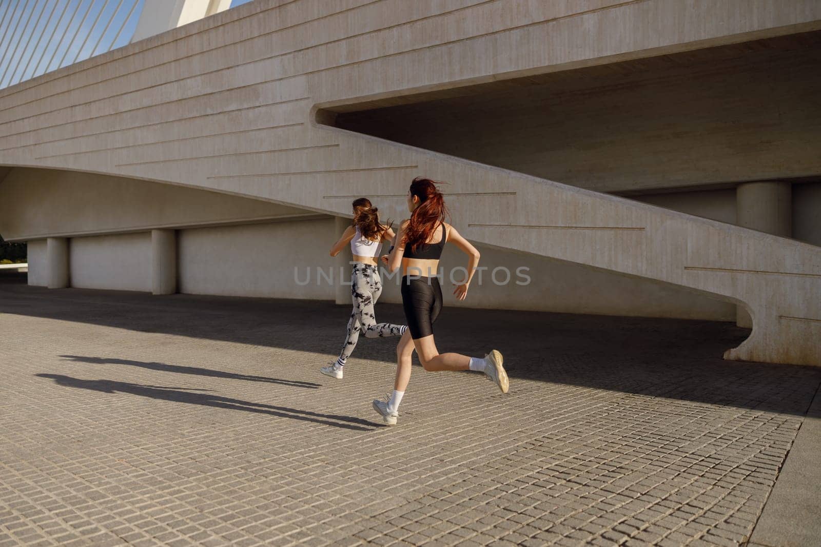 Two active women athlete running on sunny morning side by side on modern buildings background