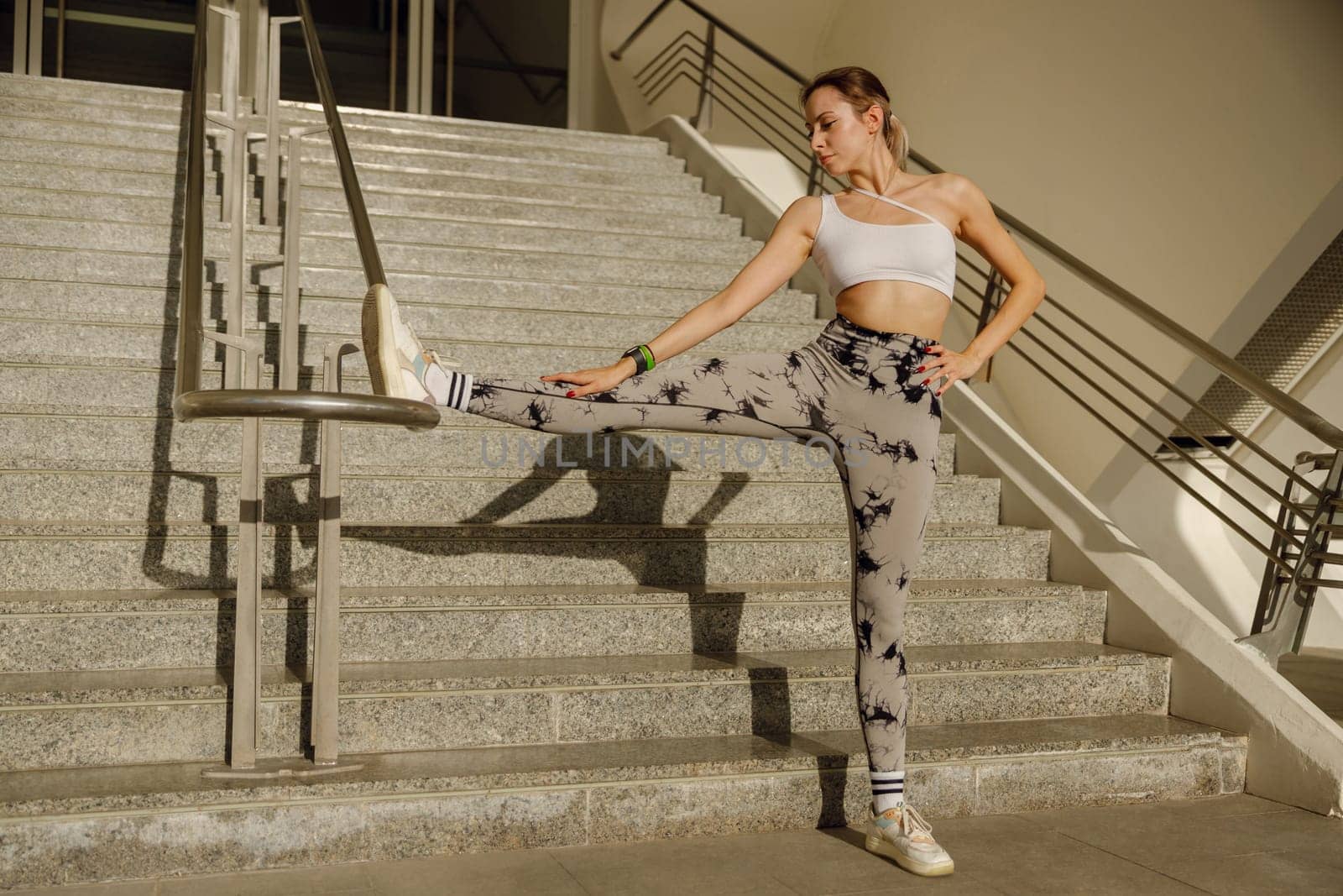 Sportswoman in tracksuit stretches legs leaning on railing on modern building background by Yaroslav_astakhov