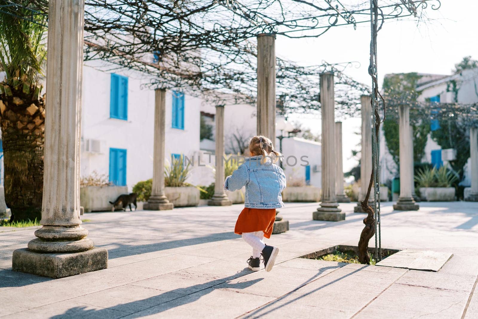 Little girl walks between pergola columns in a garden near a three-story building by Nadtochiy